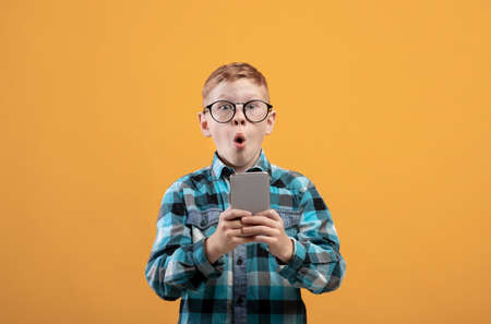 Surprised schoolboy playing game or using amazing application on smartphone over yellow studio background, copy space. Amazed ginger kid using newest mobile phone, entertainment conceptの写真素材