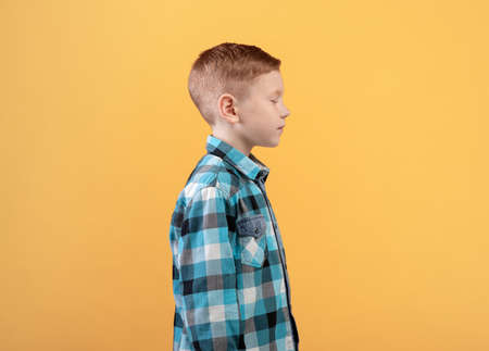 Side view of ginger boy with closed eyes posing on yellow studio background. Handsome schoolboy in casual shirt standing in profile, copy space for your text, advertisementの写真素材