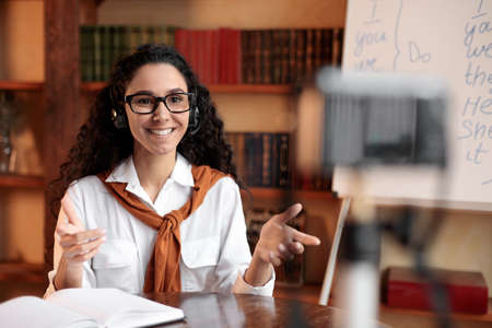 Remote Lesson. Young smiling woman wearing eyeglasses and headset giving online class, sitting at desk, explaining new theme. Female teacher recording her vlog, filming video blog at camera on tripodの写真素材