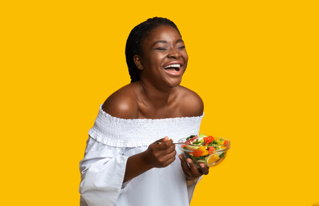 Laughing Black Woman Eating Vegetable Salad Over Yellow Background, Positive Joyful African American Lady Holding Bowl With Healthy Lunch, Enjoying Vegetarian Nutrition And Dieting, Free Spaceの写真素材