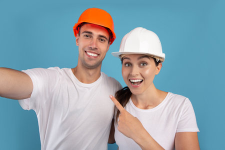 Repair Home Concept. Close up of young couple in protective helmets taking selfie together during renovation, cheerful man and woman in hard hats smiling at camera over blue background, free spaceの写真素材