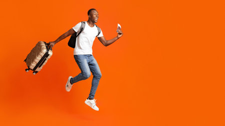 Finally vacation. Young happy black man with suitcase, backpack, passport and flight tickets jumping up orange studio background. Excited african american guy running towards copy space, panoramaの写真素材