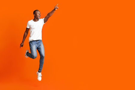 Emotional black man jumping and pointing at copy space for advertising or announcement on orange background, studio full length shot. Cheerful african american guy showing great advertisementの写真素材