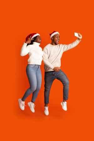 Christmas Selfie. Cheerful Black Couple In Santa Hats Taking Picture On Smartphone While Jumping Together Over Orange Background, Happy African Frineds Enjoying Celebrating Xmas Holidays, Copy Spaceの写真素材