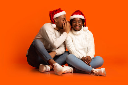 Holiday Gossips. Positive Black Guy Whispering To Girlfriends Ear, Sharing Secret Information Or Joke While They Sitting Together Over Orange Background Wearing Red Santa Hats, Copy Spaceの写真素材