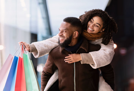 Healthy Relationship. Smiling african american man giving piggyback ride to his happy woman who holding shopping bags, couple looking at shop window, walking in the evening near centerの写真素材