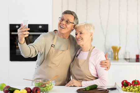 Cheerful Senior Couple Making Selfie On Cellphone Having Fun Cooking Dinner Together Standing In Modern Kitchen At Home. Elderly Spouses Enjoying Meal Preparation Using Smartphone Conceptの写真素材