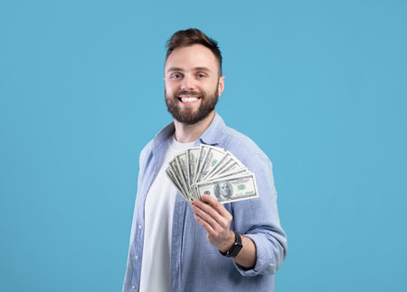 Happy young man posing with lots of money, holding dollar fan over blue studio background. Lucky millennial guy showing casino or lottery win, sharing financial success, offering profitの写真素材
