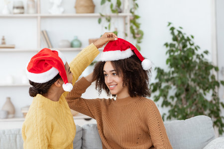 Preparing for New Year and Christmas and party at home. Cheerful african american woman in sweater puts on Santa hat to her friend and ready for holiday in interior of cozy living room, free spaceの写真素材