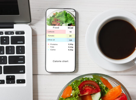 Calorie Counter Concept. Modern Smartphone With Opened Application For Counting Calories On Screen Flat Lay On Table With Coffee, Laptop And Vegetable Salad On White Tableの写真素材