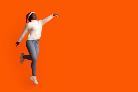 Christmas Promo. Excited Black Lady In Santa Hat Jumping And Pointing At Copy Space Over Orange Background, Cheerful African Woman Demonstrating Free Place For Your Holiday Offer Or Advertisement,の写真素材