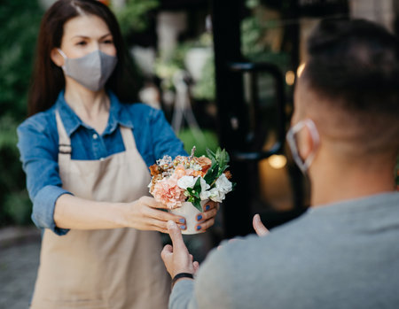 Online flower order and new normal during covid-19 quarantine and small business. Young lady owner in apron and protective mask gives bouquet to client, selective focus on composition in plant studioの写真素材