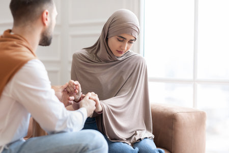 Unanswered Love. Unhappy Muslim Woman Saying No To Marriage Proposal Of Her Boyfriend, Holding Hands Sitting On Sofa At Home. Relationship Problem, Forced Marriage Concept. Selective Focusの写真素材