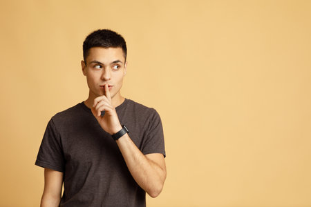 Secret, offer, interesting advertisement and gossip. Mysterious young male student with smart watch shows hand sign silence and looks towards empty space isolated on light sand background, studio shotの写真素材