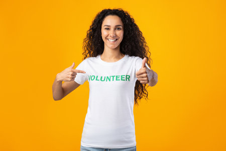 Happy Female Volunteer Gesturing Thumbs-Up Pointing At Herself Posing Standing On Yellow Studio Background, Smiling To Camera. Female Activist Showing Like Approving Volunteering Workの写真素材