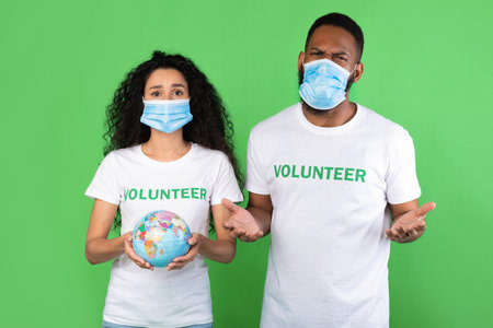 Save The Planet. Two Multicultural Volunteers Holding Earth Globe Wearing Protective Face Masks Posing Standing Over Green Studio Background. Lets Protect Our Home, Ecology And Environment Conceptの写真素材