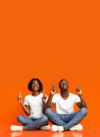 Excited african american couple sitting on floor together, pointing and looking up at copy space, orange studio background. Smiling black man and woman aiming upwards at advertisement for great dealの写真素材