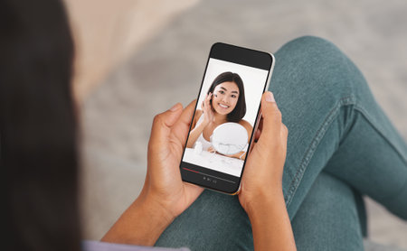 Unrecognizable black lady using smartphone to watch beauty blogger online, taking educational course on makeup. Asian influencer advising stylish cosmetics, giving web lesson remotelyの写真素材