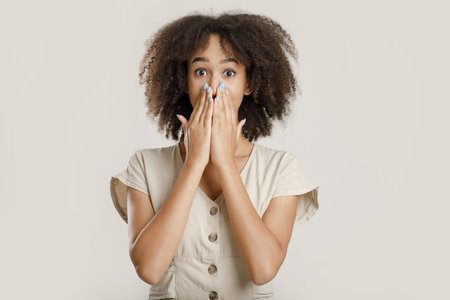 Expressing excitement, shocking news, fright and unusual proposal. Surprised millennial african american female student with curly hair in casual, closes mouth with hands, isolated on light backgroundの写真素材