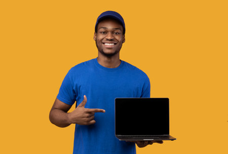 Choose Our Delivery Service. African american courier man holding laptop with blank screen for mockup, wearing blue cap and uniform, pointing at black empty monitor isolated on orange backgroundの写真素材
