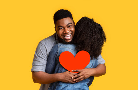 Romantic black man embracing girlfriend and holding red paper heart, making romantic surprise for...