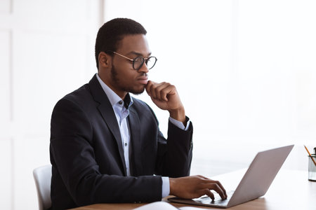 Serious african american manager in suit working with laptop in office, creating new business strategy, copy space. Young black businessman reading marketing report on computer, side viewの写真素材