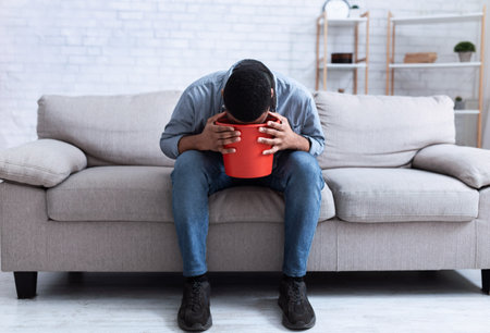 Unrecognizable African American Man Vomiting Into A Bucket Sitting On Couch At Home. Intoxication, Poisoning And Nausea, Male Health Problem. Alcoholic Addiction Concept.の写真素材