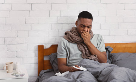 Covid-19. Sick Black Man Coughing Suffering Coronavirus Pneumonia Sitting In Bed At Home. Ill African Guy Having Inflamed Lungs Symptoms. Corona Virus, Influenza Concept. Copy Spaceの写真素材