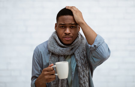 Unhappy African Man Having Headache Suffering From Cold Symptoms And Fever Touching Head Standing With Cup Of Hot Drink Over White Wall Indoors, Looking At Camera. Influenza Conceptの写真素材