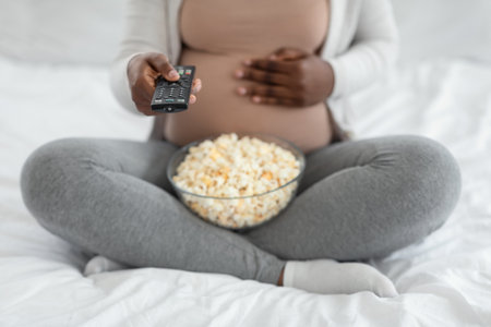 Cropped Shot Of Pregnant Woman With Popcorn And Remote Controller In Hands Relaxing At Home, Unrecognizable Expectant Mother Watching TV While Resting On Bed In Bedroom And Keeping Hand On Her Bellyの写真素材
