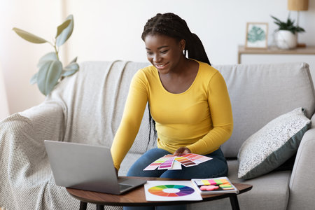 Beautiful black woman interior designer holding color swatch samples and working on laptop at home,...