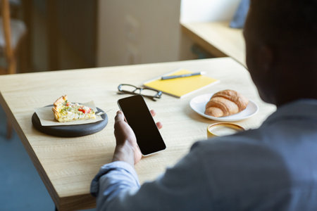 Closeup view of millennial black man holding cellphone with empty screen while having lunch at coffee shop, mockup for mobile app design. Unrecognizable guy using smartphone with space for websiteの写真素材
