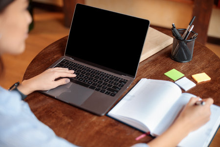 Taking Notes. Over the shoulder view of young woman sitting at desk and writing in her notebook with pen. Lady using computer with black blank empty screen, typing on keyboard, working and learningの写真素材