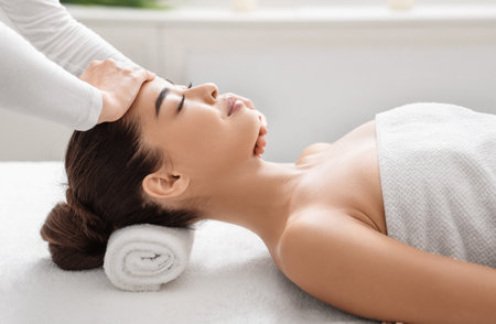 Professional therapist making relaxing head and face massage for young asian lady at spa center, beautiful korean female enjoying wellness treatments and body care, side view with copy spaceの写真素材