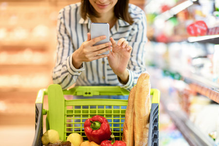 Retail And Technology Concept. Closeup cropped view of young smiling woman doing grocery shopping, using her smartphone, leaning on trolley cart. Female customer checking and comparing prices onlineの写真素材