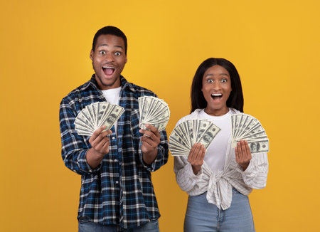 Big win, triumph together and get rich online. Happy zoomers african american male and female students with open mouths holding many dollars in hands, isolated on yellow background, empty spaceの写真素材