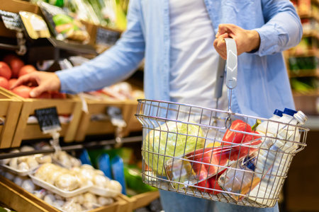 Grocery Shopping. Closeup of unrecognizable male customer holding cart basket full of food and choosing fresh eco vegetables, taking it from shelf. Young man buying healthy products in the mallの写真素材