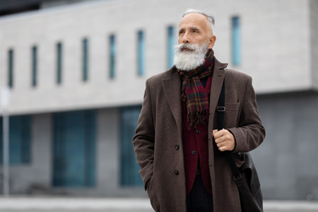 Thoughtful senior grey-haired man in stylish burgundy jacket walking by street alone, going home from job, looking at copy space. Working during retirement concept, active lifestyle of elderlyの写真素材