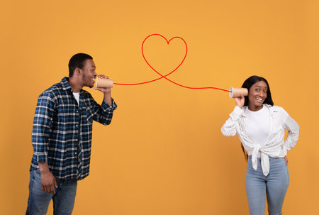 Communication, call, relationship of couple in love. Cheerful young african american student guy handing message to woman through cups and red wire, isolated on yellow background, empty spaceの写真素材