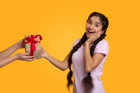 Holiday Celebration Concept. Female hands holding gift with red bow and giving box, greeting. Excited surprised indian woman taking present, looking at camera, yellow studio backgroundの写真素材