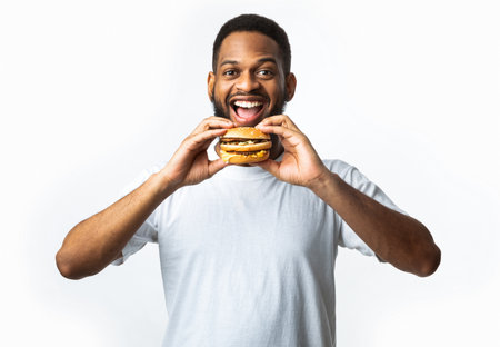 Hungry African Guy Biting Burger Eating Unhealthy Junk Food Standing Smiling To Camera Over White Background In Studio. Fast Food Overeating Habit, Unhealthy Nutrition Conceptの写真素材