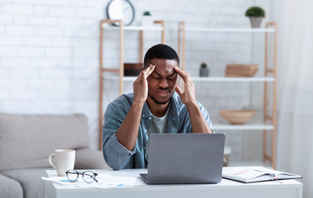 African American Businessman Having Headache Sitting At Laptop Computer Working From Home. Black Man Massaging Temples Suffering From Migraine Pain Indoor. Selective Focusの写真素材