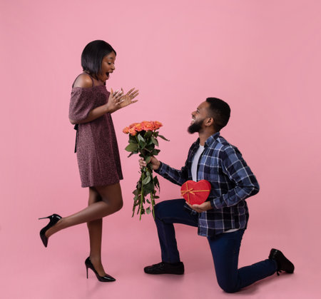 Full length portrait of romantic black guy giving his girlfriend flowers and gift for Valentines Day over pink studio background. Excited woman receiving roses and present from her boyfriendの写真素材