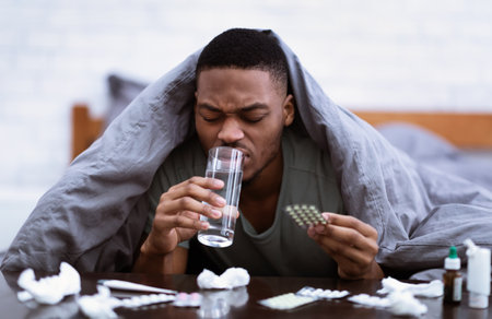 Medicine Treatment. Sick African American Man Taking Pills Drinking Water Lying In Bed At Home, With Many Medication Tablets And Painkilling Capsules. Healthcare, Medical Drugs Prescriptionの写真素材