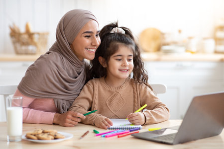 Smiling Muslim Mother And Her Little Daughter Using Laptop In Kitchen Together, Loving Islamic Mom Helping Girl With Online Education And Development Activities, Looking At Computer Screen And Smilingの写真素材