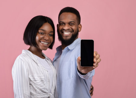 Romantic dating app. Affectionate black couple showing mobile phone with empty screen on pink studio background, space for mockup. Happy woman and her boyfriend advertising your website designの写真素材
