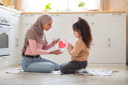 Happy Mothers Day. Little girl greeting her muslim mom in hijab with gift card with big red heart at home, surprised islamic mommy and cute daughter sitting together on floor in kitchen, copy spaceの写真素材
