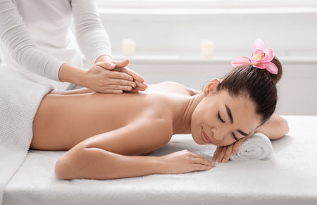 Beautiful Asian Woman Lying On Spa Bed, Getting Back Massage Treatment From Professional Masseur In Beauty Salon, Relaxed Korean Lady Enjoying Body Care And Self-Pampering, Side View With Free Spaceの写真素材