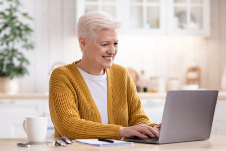 Happy grey-haired senior woman sitting in kitchen and using laptop, typing on keyboard, copy space....