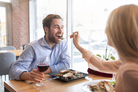 Positive young couple having romantic festive dinner at cafe, celebrating birthday, feeding each other, drinking wine. Lovely lady giving her boyfriend yummy fresh salad at coffee shopの写真素材
